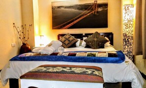 A hotel room with a bed, a nightstand, a vase with branches, a framed picture of a bridge, and a curtain with text.