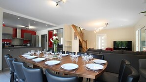 Un comedor moderno con una mesa larga dispuesta para seis comensales, una cocina al fondo, una escalera y una sala de estar con sofá y televisor.
