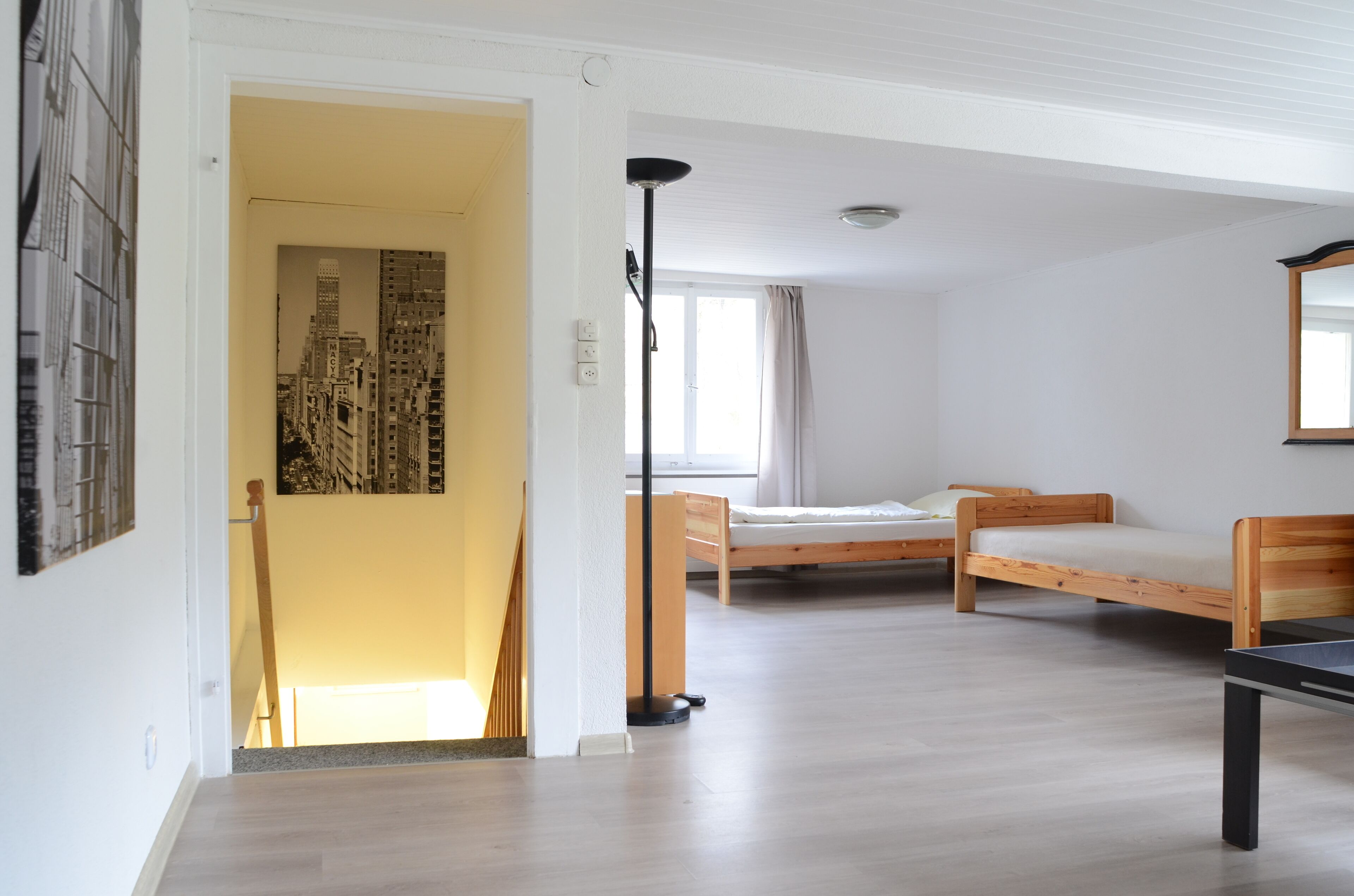 Une chambre avec deux lits en bois, une lampe sur pied et un tableau représentant un paysage urbain, encadré et accroché au mur.