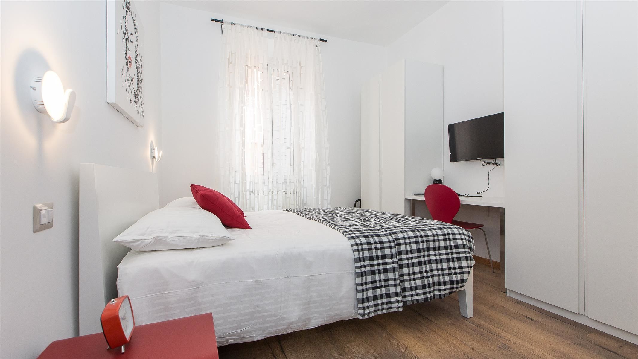 A bedroom with a bed, a desk, a TV, and a window with curtains.