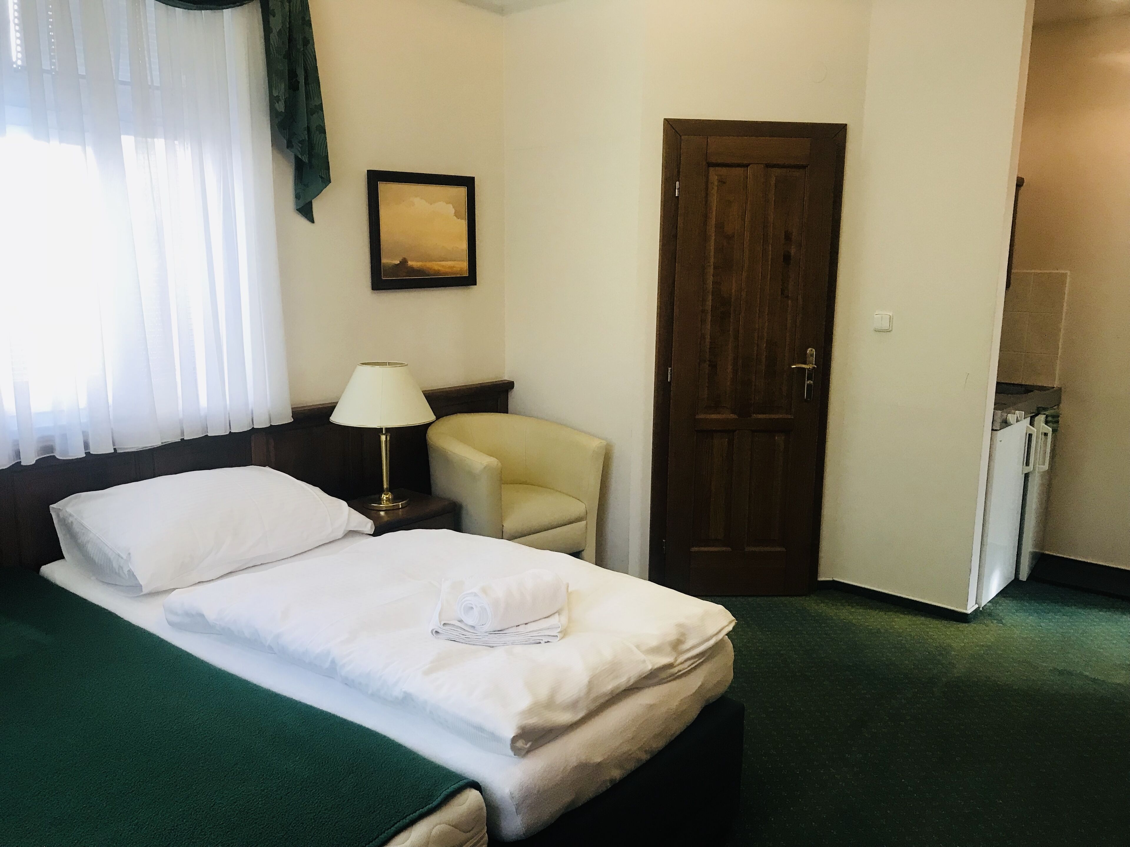 Una cama bien hecha con sábanas blancas, una lámpara de noche, una silla, una puerta y un pequeño armario.