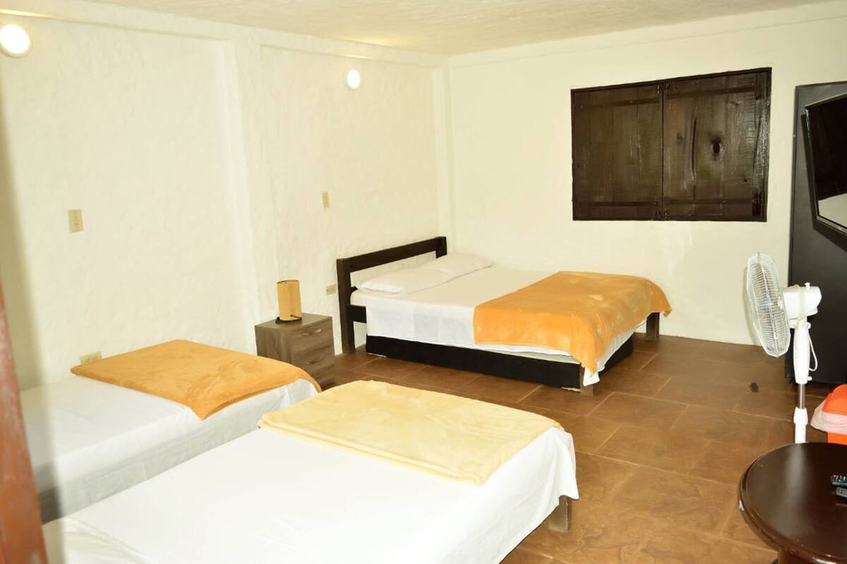 A room with two beds, a bedside table with a lamp, a wall-mounted fan, and a dark wooden panel on the wall.