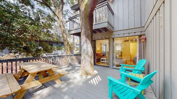 A wooden deck with picnic tables and turquoise chairs, a tree, and a building with a balcony.