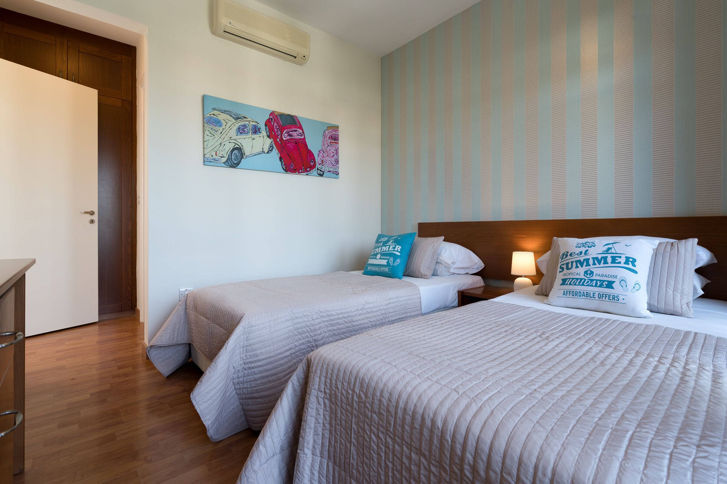 A hotel room with two beds, a wooden floor, a striped wall, and a painting of cars.