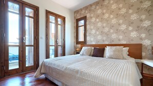 Ein Schlafzimmer mit einem großen Bett, Holzboden, blumigem Wandpapier und einem Fenster mit Fensterläden.