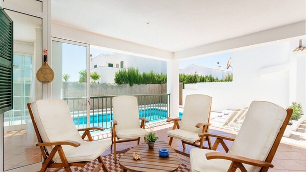 Eine sonnendurchflutete Terrasse mit vier weißen, gepolsterten Stühlen, einem runden Holztisch und Blick auf einen Swimmingpool und viel Grün.