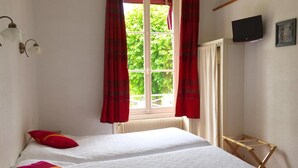 A bedroom with two beds, red curtains, a window with a view of greenery, a radiator, a TV, and a wooden drying rack. - Hotel le Vendangerot (Rully)