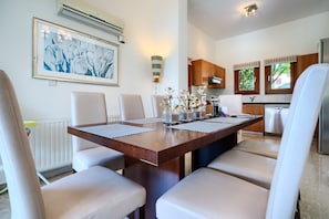 Un comedor con una mesa de madera y sillas blancas, un cuadro enmarcado en la pared y una cocina al fondo.