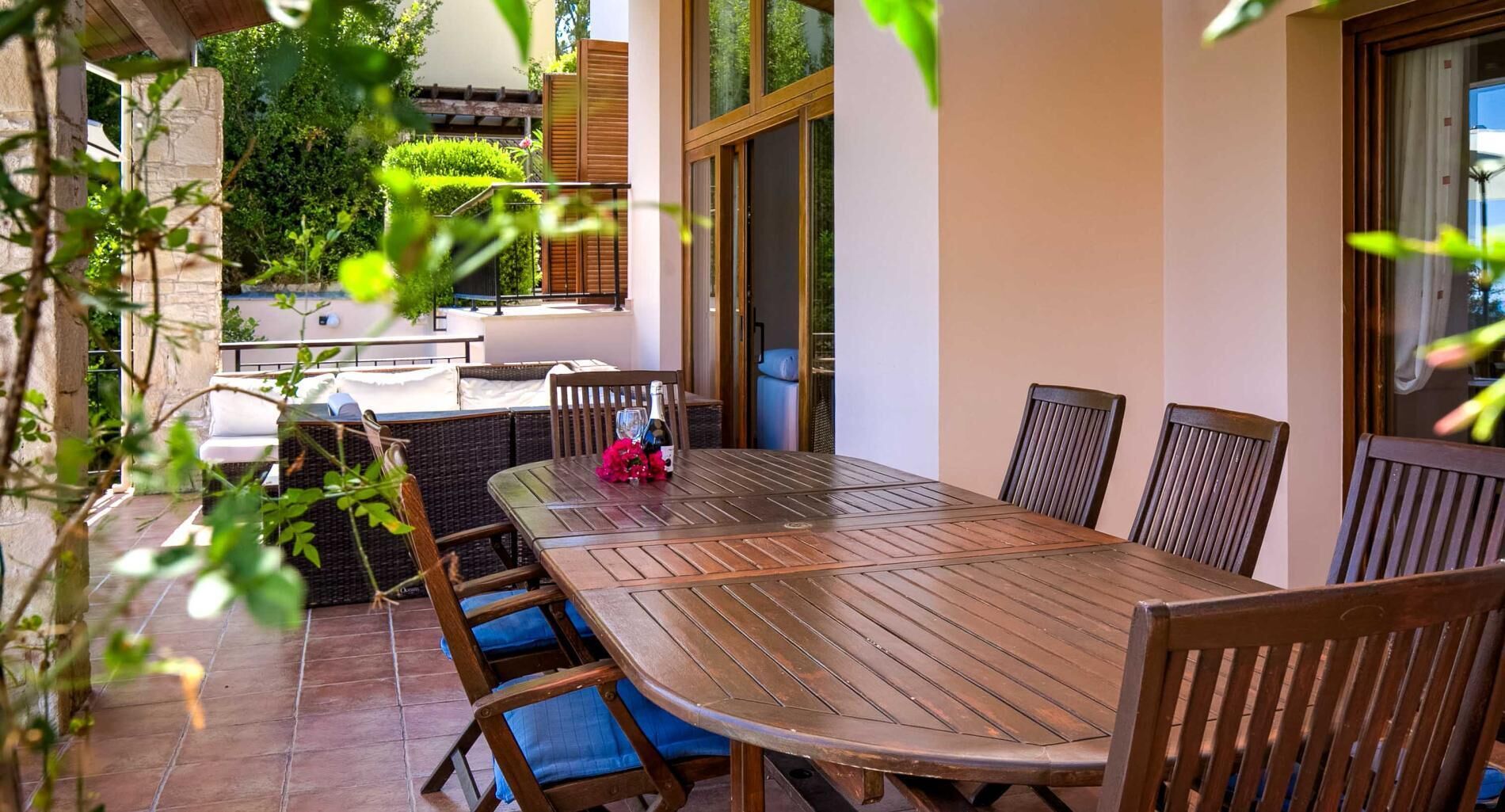 A patio with a wooden table and chairs, surrounded by greenery and a stone wall.