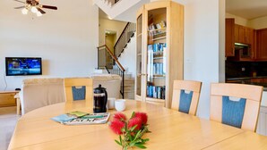 Ein modernes Wohnzimmer mit einem Holzesstisch, einer Kaffeemaschine und einer Zeitschrift. Eine Treppe führt zu einer anderen Etage, es gibt eine Küchenzeile und ein Bücherregal mit Büchern.