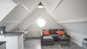 Een modern loft appartement met een schuine plafond, een kitchenette, een grijze bank met oranje kussens en een rond raam dat natuurlijk licht binnenlaat.