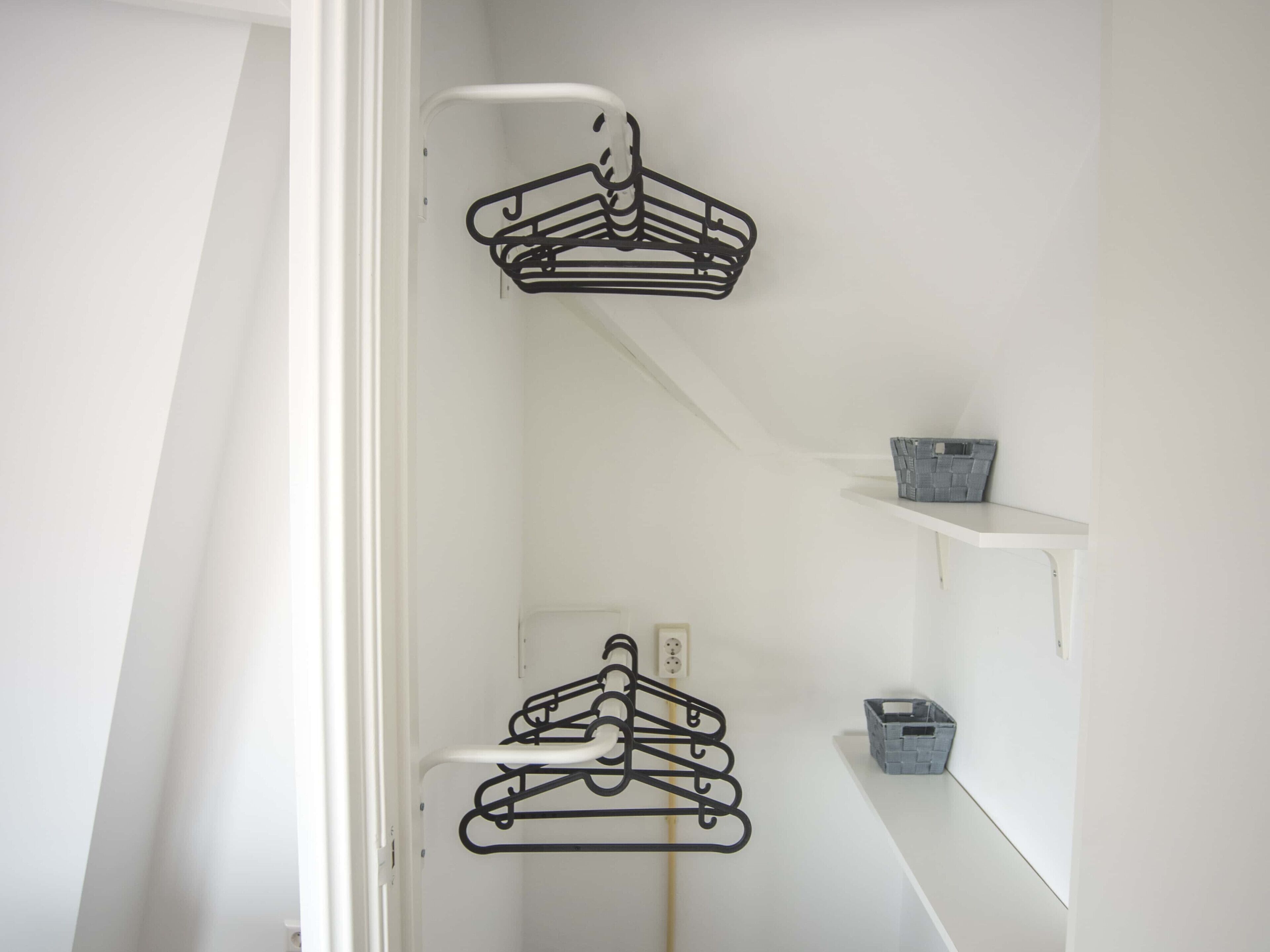 A room with white walls, a white door, and a white ceiling. There are two black metal hangers hanging on a white metal rod. There is a small gray basket on a white shelf.