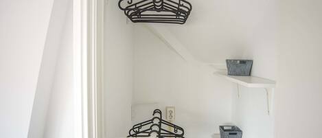 A room with white walls, a white door, and a white ceiling. There are two black metal hangers hanging on a white metal rod. There is a small gray basket on a white shelf.