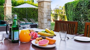 An outdoor dining area with a table set for breakfast, including a bottle of green juice, a pitcher of orange juice, a plate with grapefruit, bananas, and pastries, and a glass of water. - Apartment DC01 - Kalypso (Kouklia)