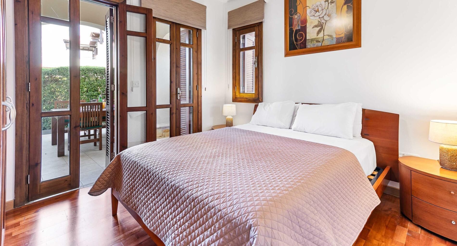 A bedroom with a large bed, wooden floor, a nightstand with a lamp, a painting on the wall, and a sliding glass door leading to an outdoor area with a table and chairs.