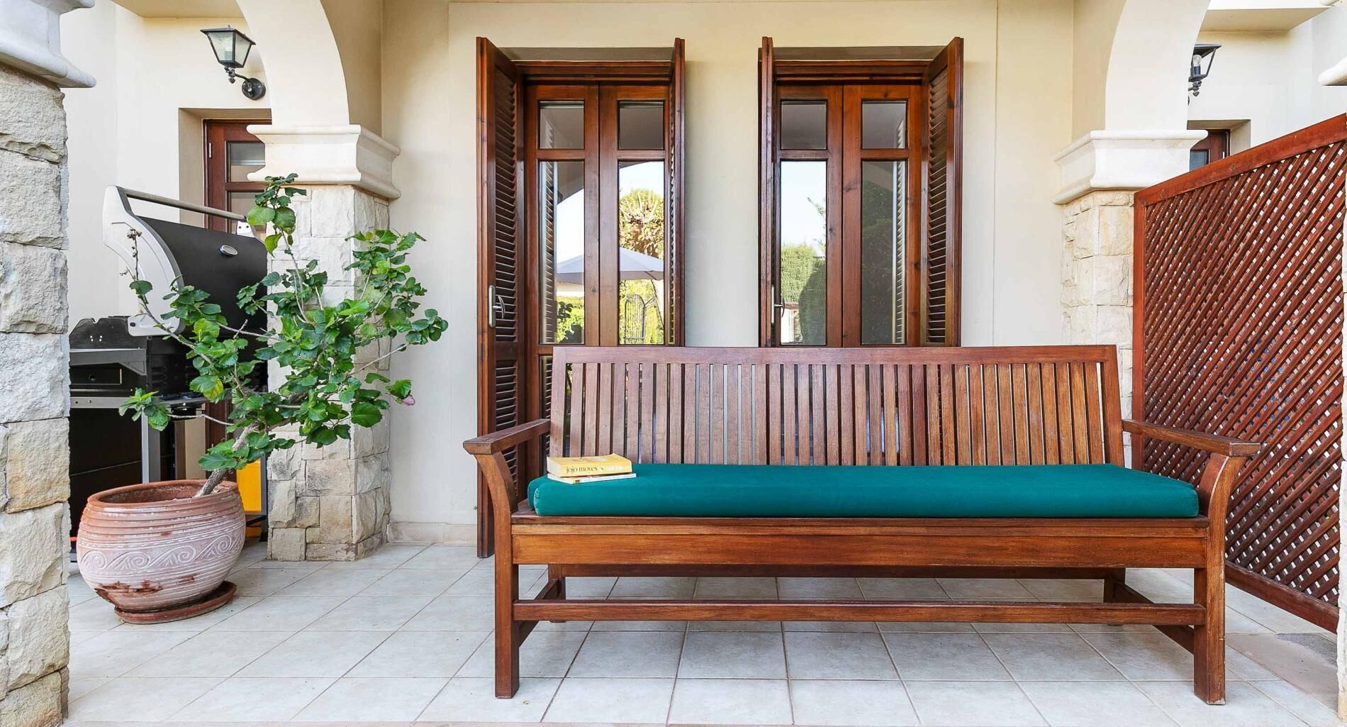 A wooden bench with a green cushion, a potted plant, and a grill outside a building with stone walls and wooden shutters.