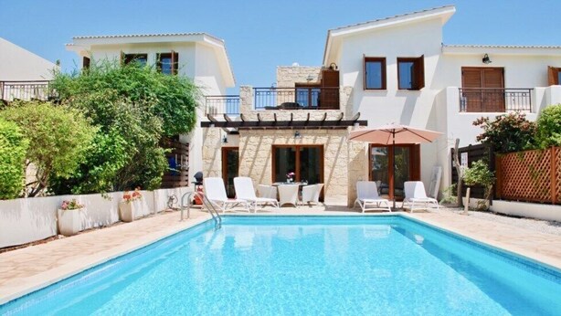 Uma área de piscina com espreguiçadeiras e uma mesa, cercada por um edifício branco com detalhes em pedra e uma varanda.