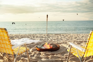 A beach scene with two yellow and white striped beach chairs facing a fire pit on the sand. - Hotel Corduroy (Montauk)