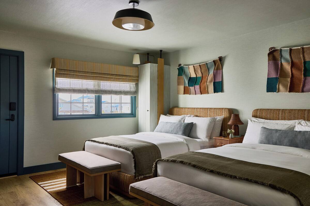 Een hotelkamer met twee bedden, een tafeltje, een bankje, een raam met zonwering en een wandlamp.
