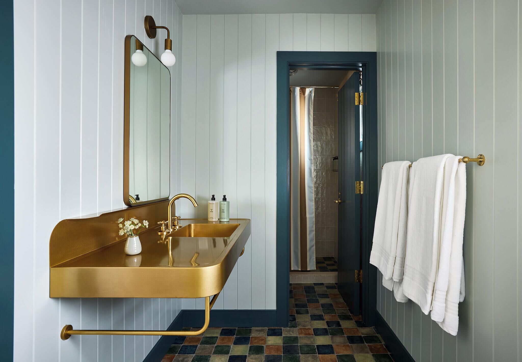 A bathroom with a gold sink, a mirror with lights, a towel rack, and a shower area.