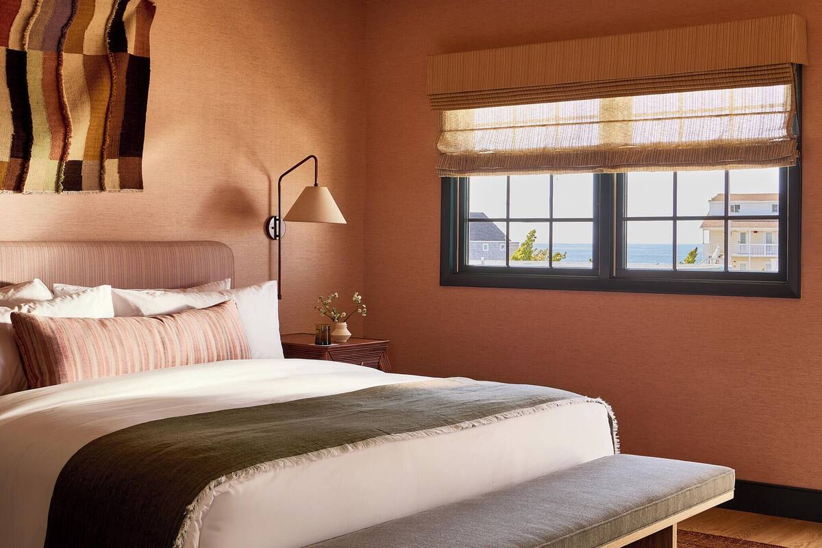 Een slaapkamer met een bed, bedlampje, raam met uitzicht en een wanddecoratie.