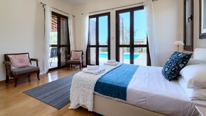 Ein Schlafzimmer mit einem großen Bett, einem Sessel, einem Fenster mit Vorhängen und Blick auf einen Pool.