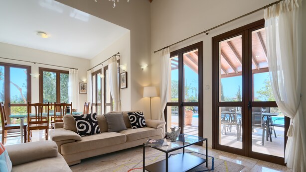 Ein geräumiges Wohnzimmer mit einem beige farbenen Sofa, einem Glastisch und einem farbenfrohen Teppich. Große Fenster und eine Terrasse mit Gartenmöbeln sind zu sehen.