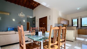 Ein Essbereich mit Glastisch und Holzsesseln, einer Couch, einer Pendelleuchte und einer Küche mit Holzschränken und einem Fenster.