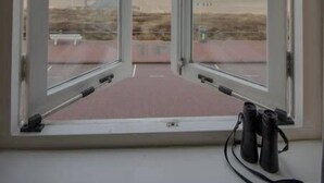 Una ventana con binoculares en el alféizar, con vistas a un paisaje llano y estructuras distantes.