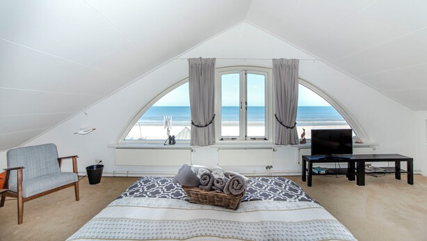 Un dormitorio con una cama grande, una silla, un televisor y un ventanal con cortinas.