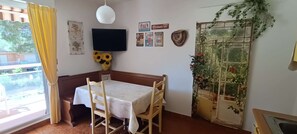 Quarto com mesa, cadeiras, televisão na parede e janela com cortinas.