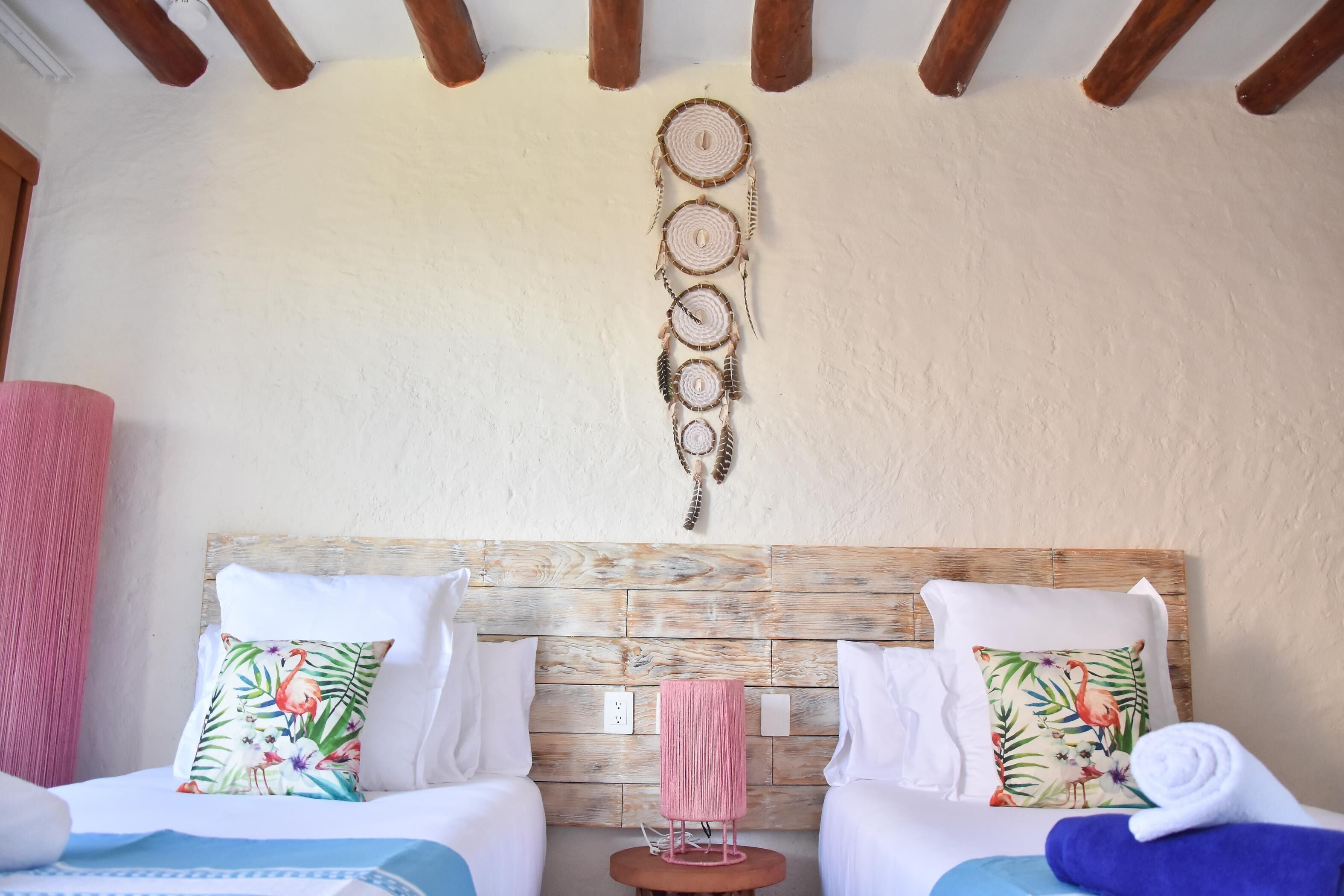 A room with two beds, each with a floral pillow, a wooden headboard, and a small round table with a pink lamp.