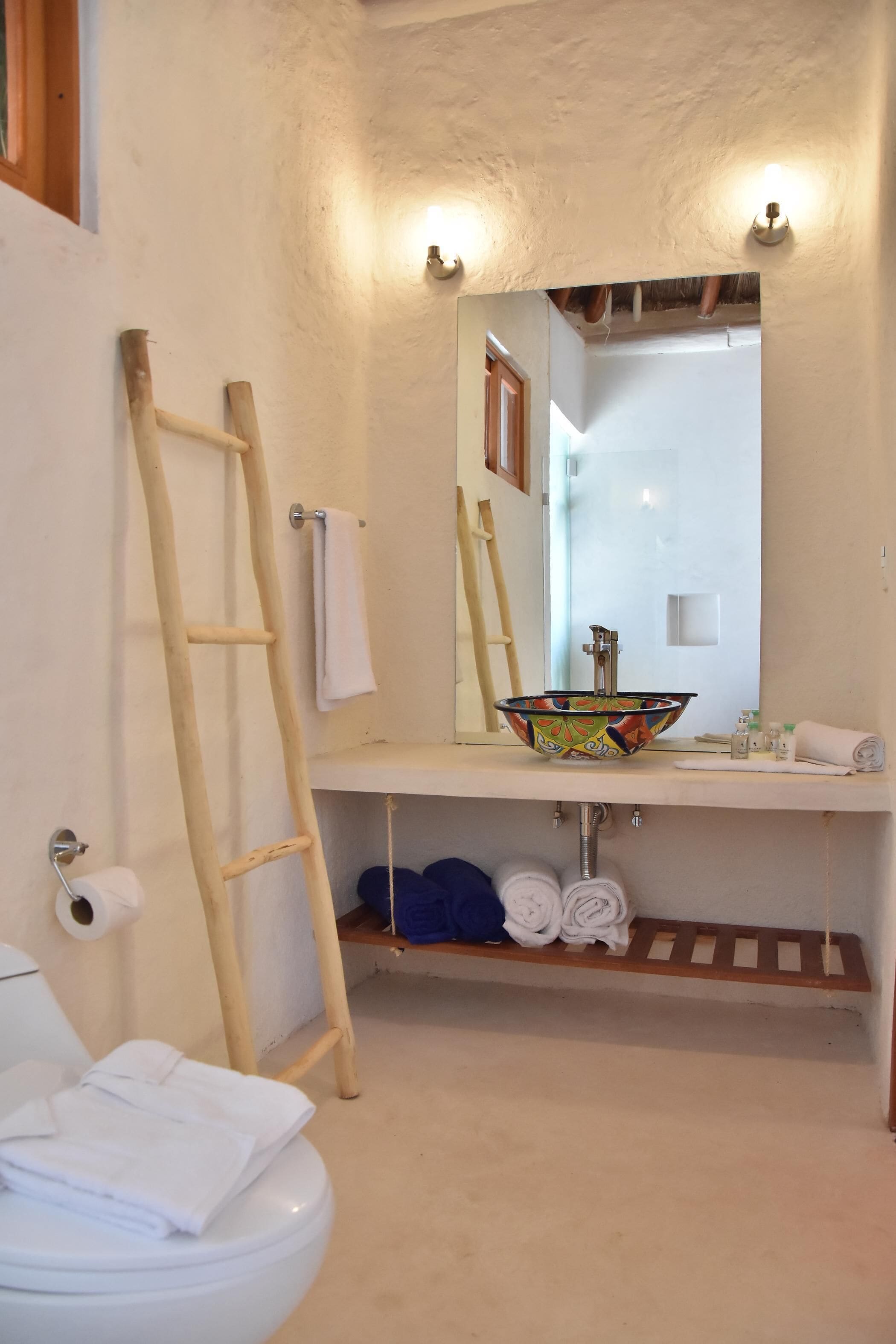 Un baño con una escalera de madera, un inodoro blanco, un lavamanos con un cuenco de colores y un espejo.