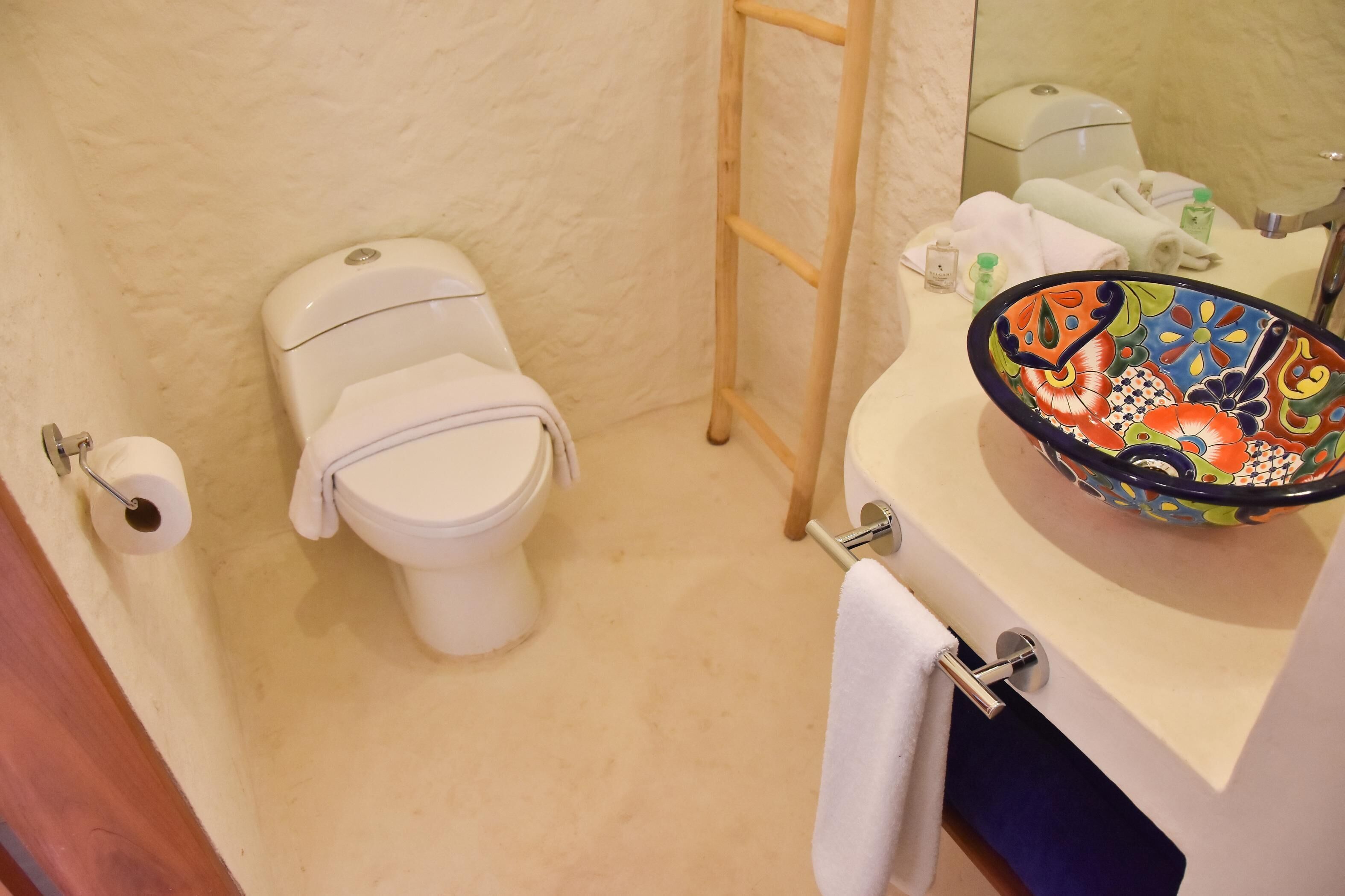 Un baño con inodoro, toallero, lavamanos y un recipiente de colores, y un espejo.
