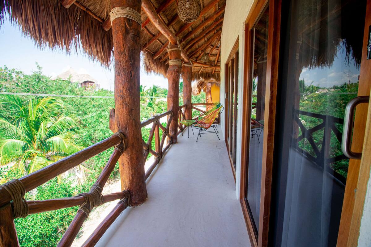 A wooden balcony with a thatched roof, overlooking lush greenery.