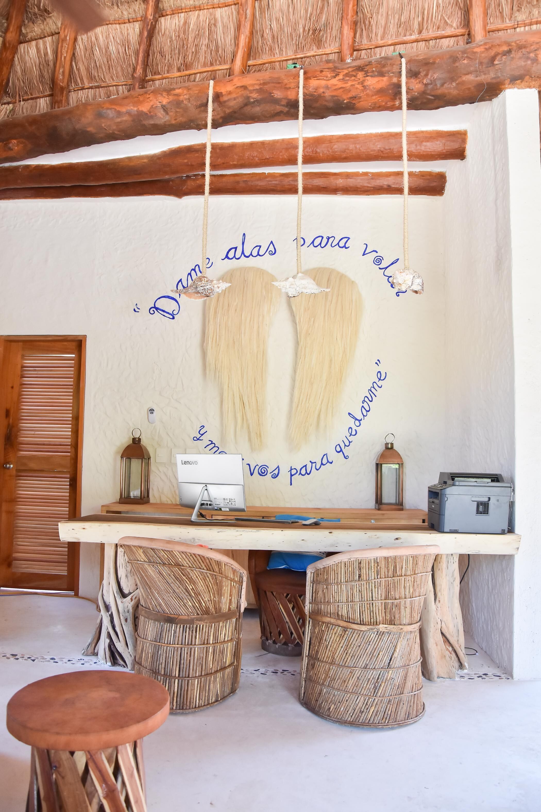 Un interior rústico con techo de madera, pared blanca con texto decorativo, un escritorio con computadora y dos taburetes de mimbre.