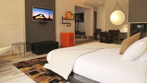 A modern hotel room with a large bed, a TV mounted on the wall, a desk with a chair, and a small seating area with a chair and a table. - Le Stanislas Maison d'Hôtes (Abidjan)