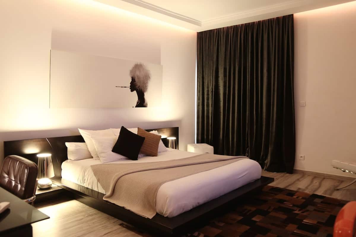 A modern hotel room with a large bed, a desk with a lamp, a chair, and a large window with dark curtains.