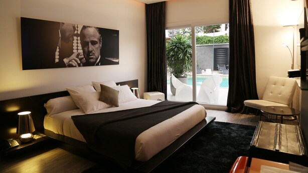 A modern hotel room with a large bed, a desk, a chair, a large window with a view of a pool and greenery, and a painting on the wall. - Le Stanislas Maison d'Hôtes (Abidjan)