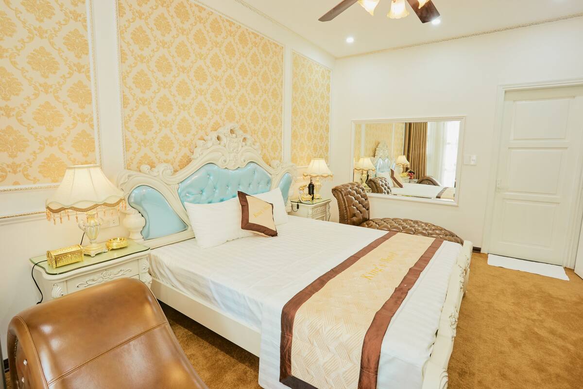A hotel room with a large bed, a sitting area with a chair, a small table with a lamp, and a mirror on the wall.