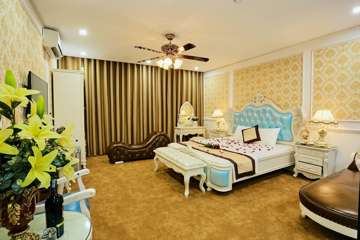 A spacious bedroom with a large bed, a ceiling fan, a bedside table with a lamp, a nightstand, and a decorative mirror.