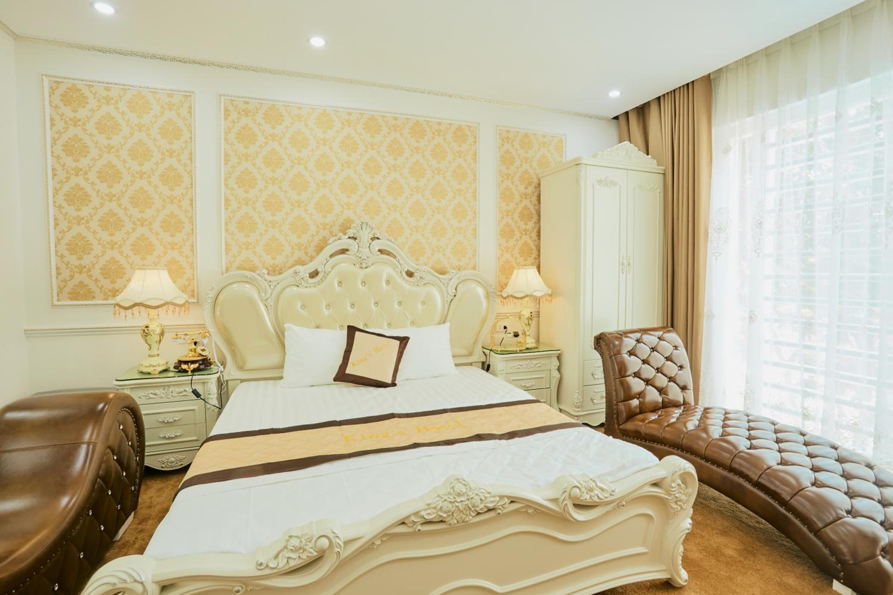A hotel room with a large bed, a brown leather armchair, a white wardrobe, and a large window with curtains.