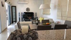 A modern living room with a flat-screen TV, a dining area with a granite countertop, and a large window with blinds. - Marina Thea Villa No. 11 (Coral Bay)