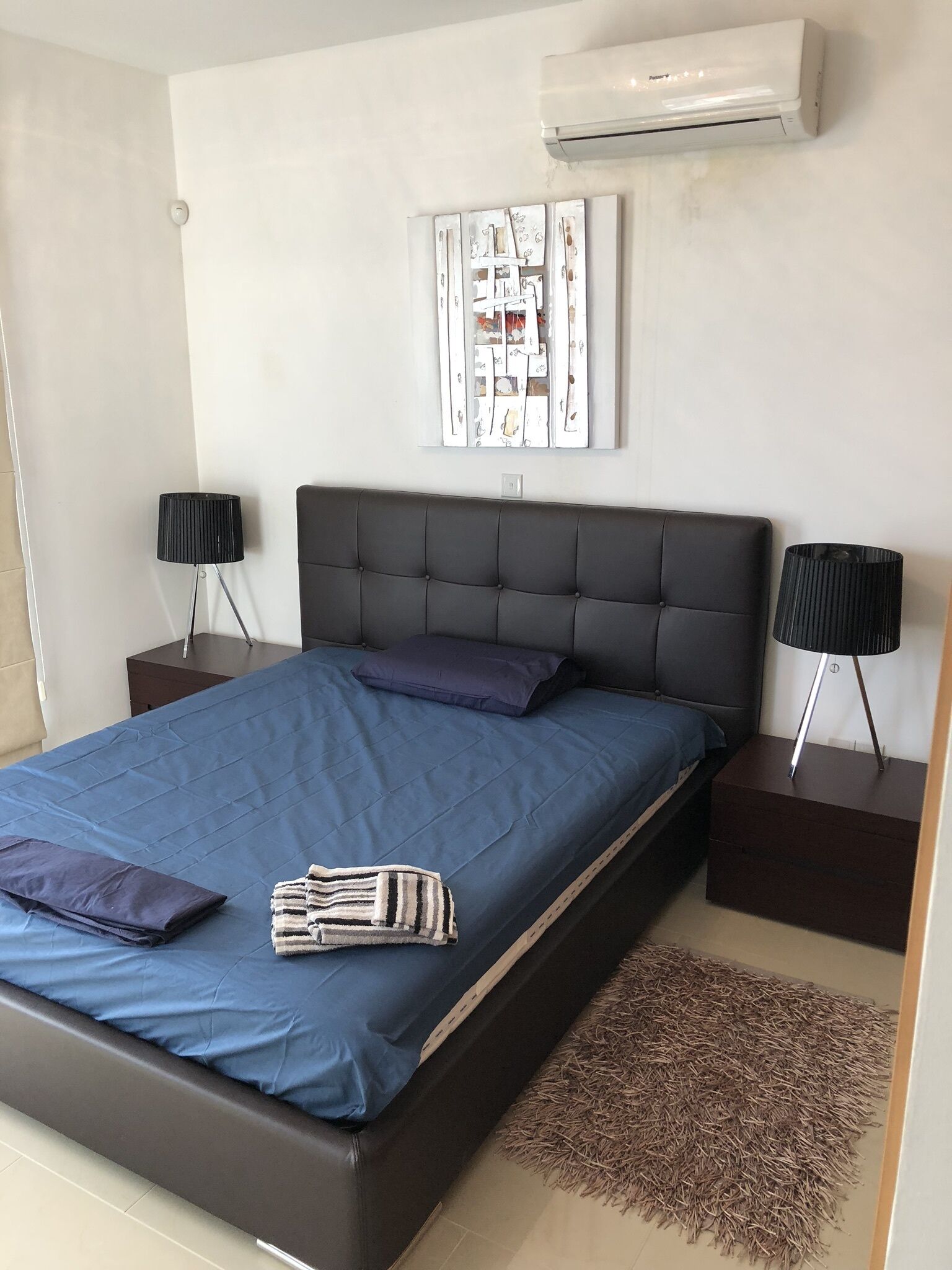 Un dormitori modern amb un llit gran, dues mesetes de nit, un aire condicionat fixat a la paret i una peça decorativa sobre el llit.