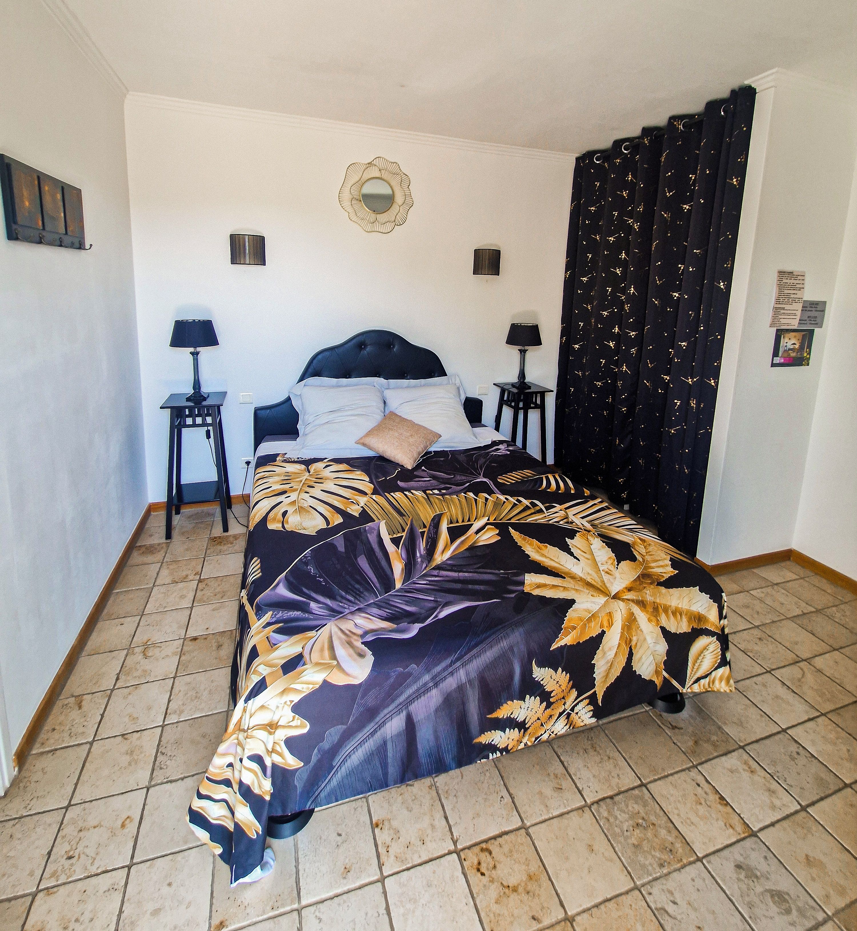 Una cama con una colcha estampada con motivos de hojas, dos mesitas de noche con lámparas, un espejo en la pared y una cortina negra con un diseño dorado.