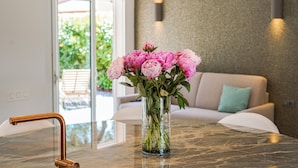 Een marmeren tafel met een heldere vaas met roze bloemen, een moderne kraan en een bank met een blauwgroene kussen op de achtergrond.