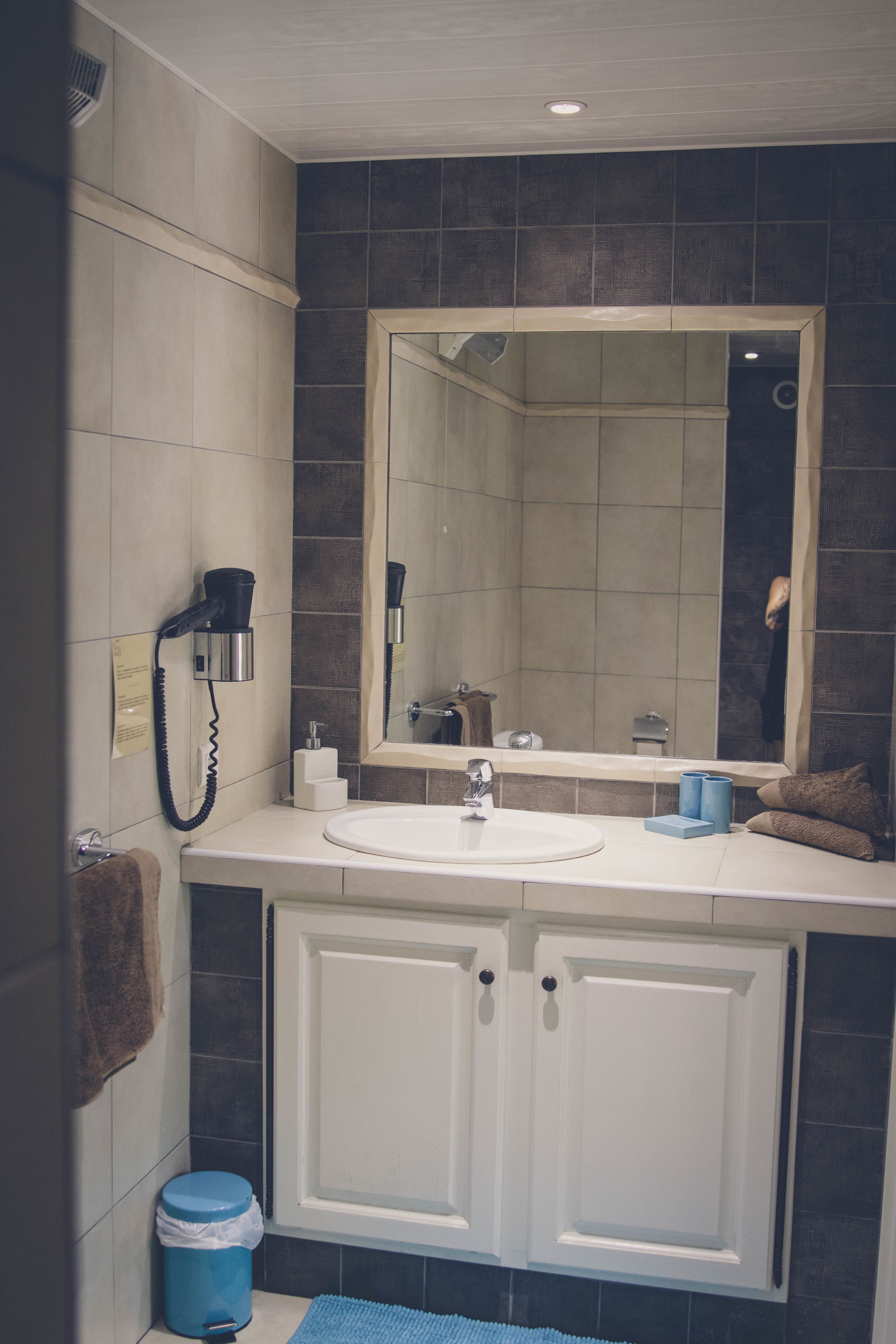 Un baño con lavamanos blanco, un espejo grande, secador de pelo y un cubo de basura azul.