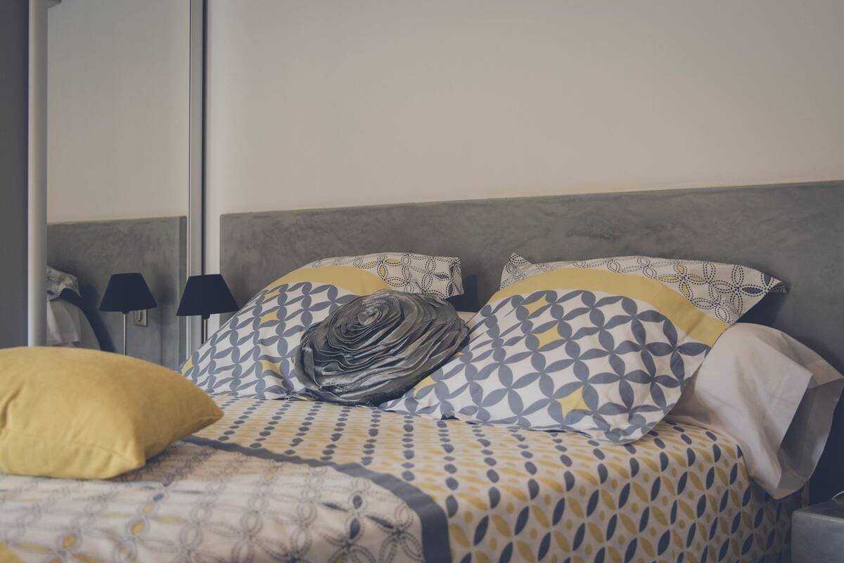 Een net opgemaakt bed met behangpatroon en kussens, een bedlampje en een spiegel aan de muur.
