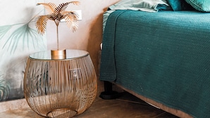 Een bed met een blauwgroene spreidek, een rond geweven bijzettafeltje met een decoratieve plant, en een muur met een bladmotief.