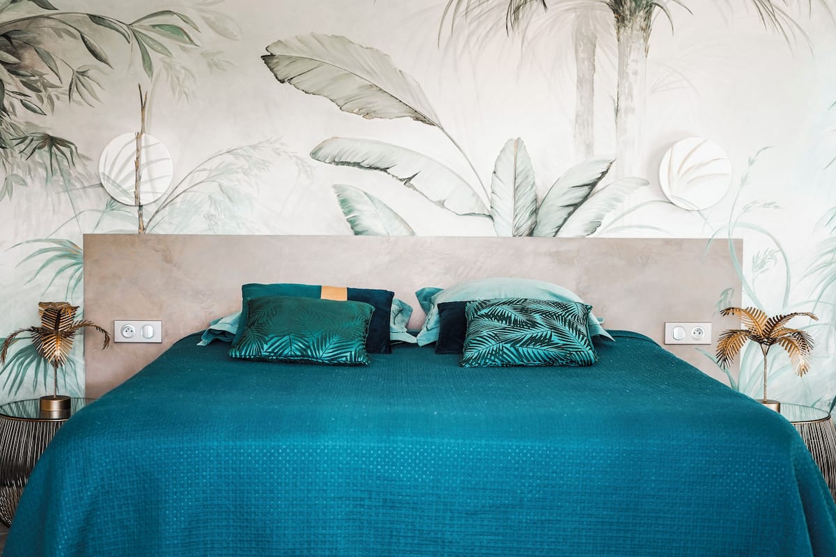 Een bed met een blauwgroene dekbedovertrek, twee kussens met een bladpatroon, en een muur met een grote plantenmuurschildering.